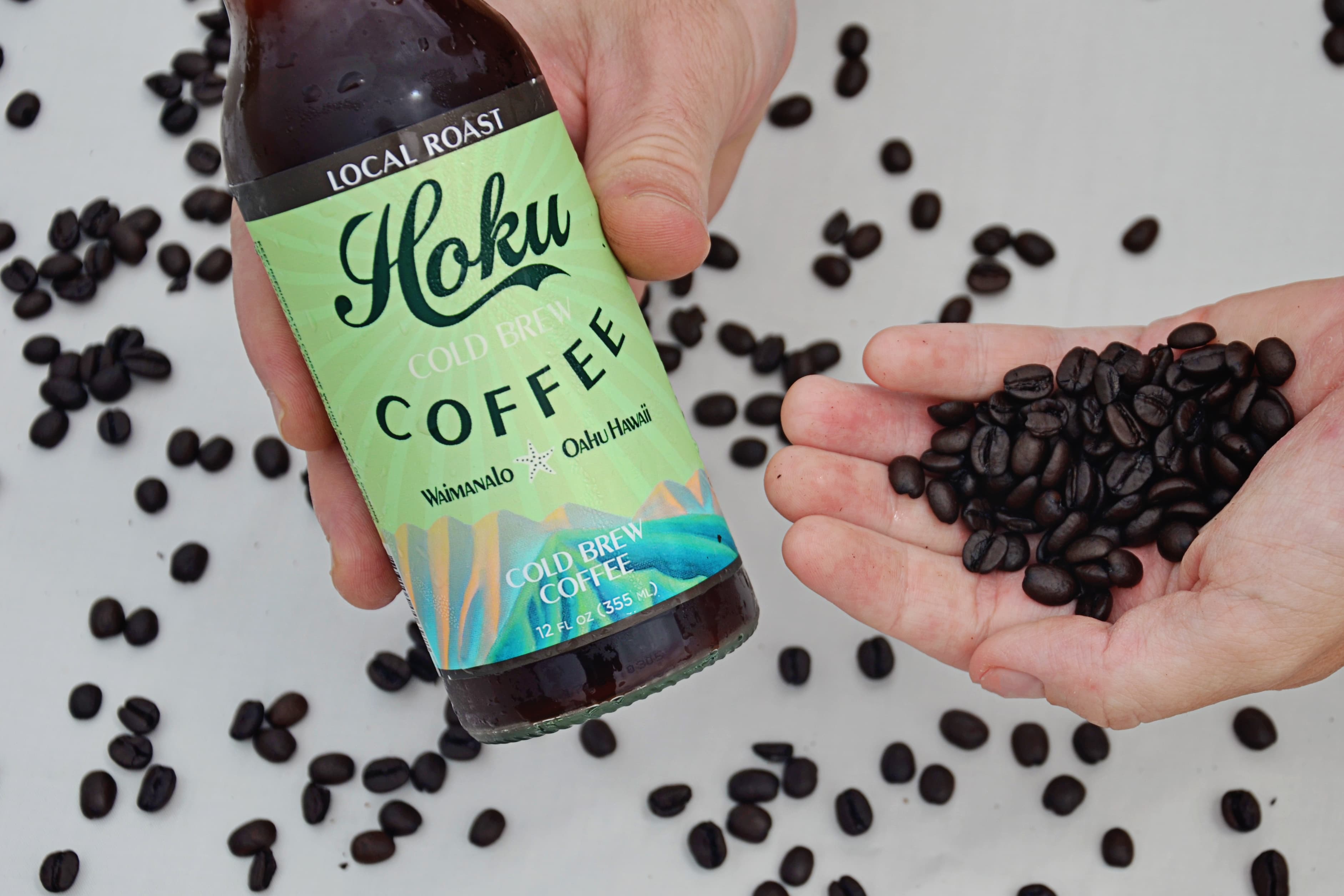Hand holding a Hoku Local Roast bottle beside a hand of roasted beans